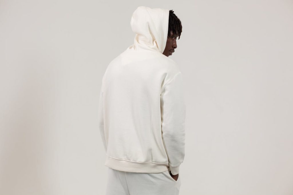 Back view serious African American male wearing white comfy hoodie and pants standing with hands in pockets against light studio wall and looking away