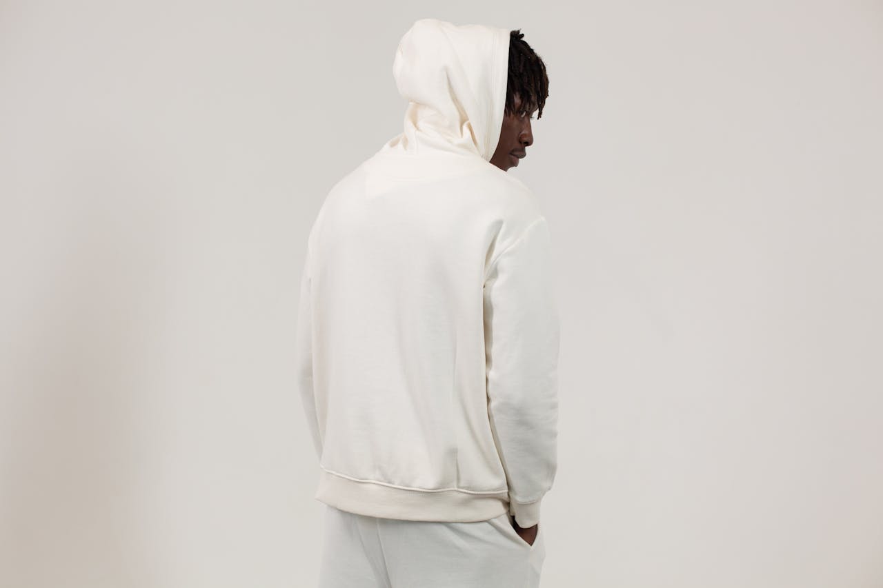 Back view serious African American male wearing white comfy hoodie and pants standing with hands in pockets against light studio wall and looking away